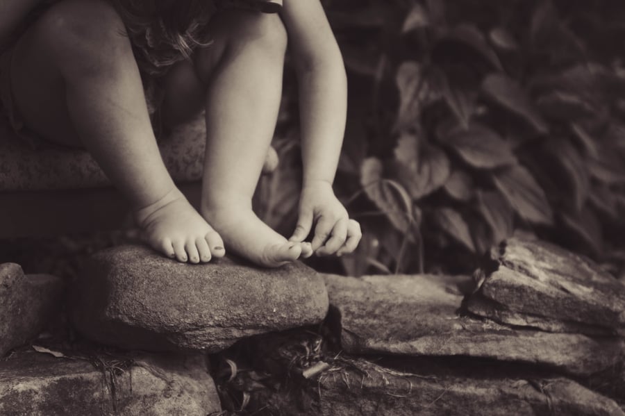 Sweeney Foot and Ankle FAQs About Plantar Warts in Children Black and White Photo of Young Child Picking at Toes and Sitting On Rocks